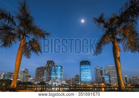 Lake With Palm Trees In City Park Under Skyscrapers At Night. Benjakiti Park In Bangkok, Thailand