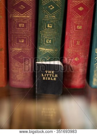 A Small Holy Book Of The Bible Stands On A Shelf Near Large Multi-colored Books.