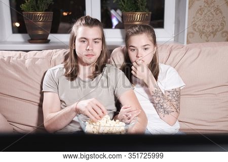 Portrait Of A Long-haired Guy And Girl With Popcorn In Their Hands On The Couch Watching Tv. Family 