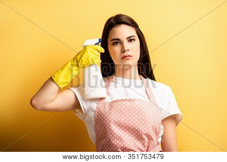 Serious Housewife In Apron And Rubber Gloves Imitating Self-killing With Spray Bottle On Yellow Back