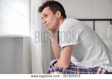 Young Man In The White Shirt And Pajama Sitting On A Bed And Looking At The Wondow. Reflexing About 