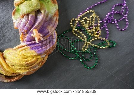 Flay Lay Of A Festive Mardi Gras King Cake With Purple, Gold And Green Beads With A Small Plastic Ba