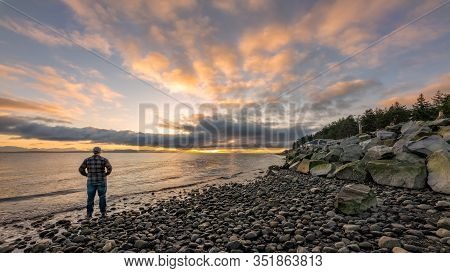 Man Standing Along Shore Looking Into Dramatic Beautiful Sunrise Along Rocky Beach.