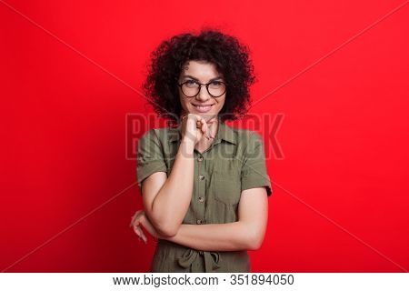 Charming Caucasian Woman With Curly Black Hair Is Touching Her Chin While Looking Through Glasses An