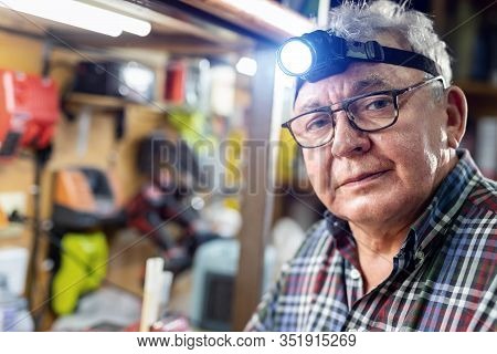 Senior Craftsman Portrait With Headlamp Led Torch Sitting In Workhop With Tools And Equipment On Bac