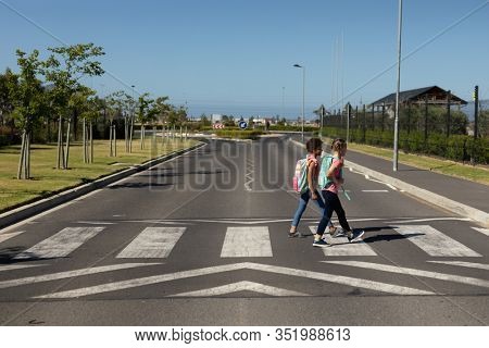 Side view of a Caucasian and an African American schoolgirl crossing the road on a pedestrian crossing on their way to elementary school on a sunny day