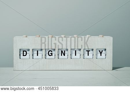 the word dignity in a rustic wooden split flap board, on a wooden surface in front of a pale gray wall