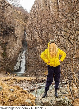 A Woman In A Yellow Jacket With Hair Instead Of A Face Stands Against The Backdrop Of Rocks And A Wa