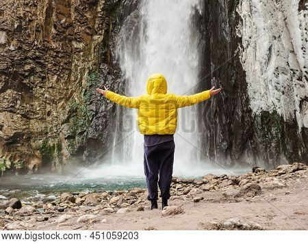 A Woman In A Yellow Jacket Stands With Her Hands Apart, Looking At A Stormy Ice Waterfall Flowing Do