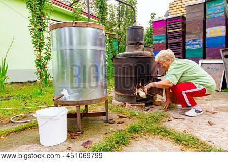 Grandma Throws Dry Branches Into The Firebox Of A Homemade Distillery Making Moonshine Schnapps, Alc