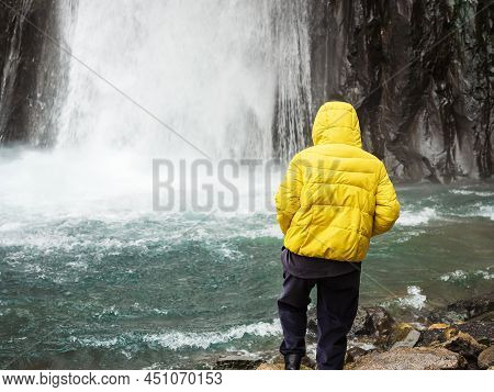 A Woman Takes A Step Into A Turquoise Mountain River Into Which A Powerful Icy Stream Of A Waterfall