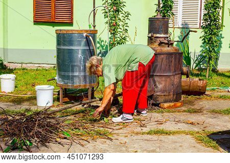 Grandma Throws Dry Branches Into The Firebox Of A Homemade Distillery Making Moonshine Schnapps, Alc