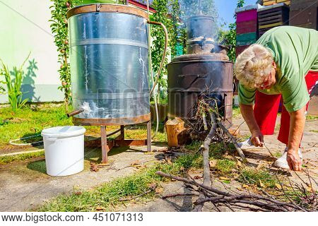 Grandma Throws Dry Branches Into The Firebox Of A Homemade Distillery Making Moonshine Schnapps, Alc