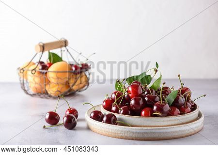 Fresh Cherries In Two Light Ceramic Plates And Apricots In A Wicker Iron Basket On A Gray Concrete A