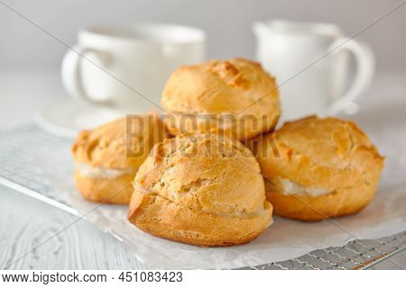 Homemade Profiteroles On A Metal Grid And White Paper On A White Wooden Background. Behind Are A Tea