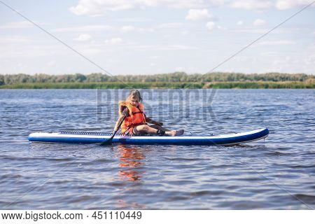 Female Kid Sitting On Sup Board By Herself On Lake Holding Oar In Right Hand And Looking At Rippled 