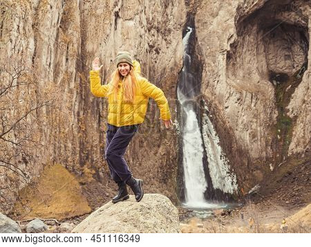 A Cheerful Girl Stands On A Stone In A Pose Against The Backdrop Of Steep Caucasian Rocks And A Wate