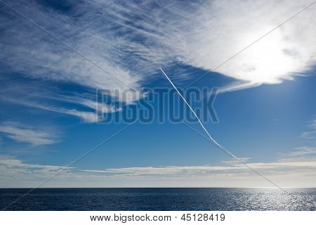 A sharp jet contrail through blue sky entering a high cloud area over a blue sea