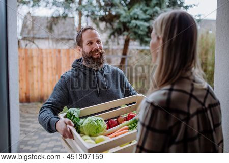Mature Man Holding Crate With Vegetales And Fruit And Delivering It To Woman Standing At Doorway.
