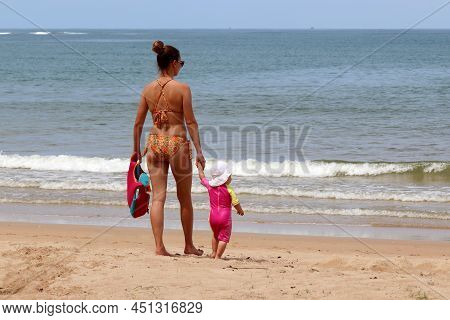 Young Mom Leading Kid To The Sea. Slim Woman In Bikini With Little Child On A Sandy Beach