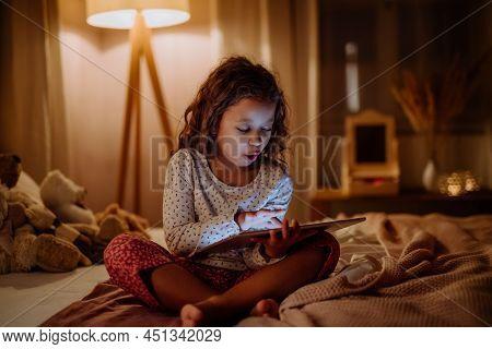 Happy Little Multiracial Girl Using Tablet On Bed In Evening At Home.