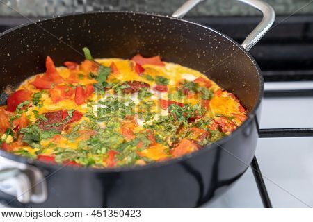 Omelet With Vegetables And Herbs In Black Frying Pan On White Stove. Side View