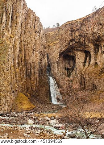 In The Steep Cliffs With Caves, A Powerful Stream Of A Waterfall Flows, Falling Into The River Of Th