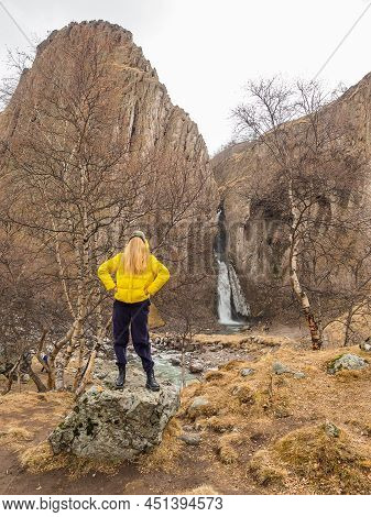 A Woman With Her Face Hidden Under Her Hair Stands On A Rock In A Rocky Gorge With A Waterfall In Th