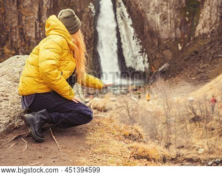 A Woman In A Yellow Jacket Squatting On The Background Of Autumn Nature Put Her Hand To A Waterfall 