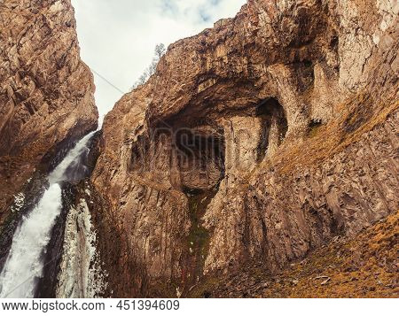 A Powerful Waterfall Flows Down From Steep Cliffs Against A Cloudy Sky. Bottom View