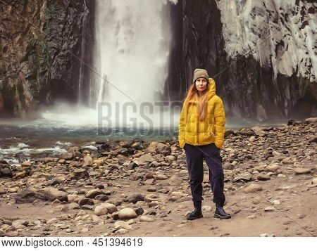 The Girl Stands Thinking Against The Backdrop Of A Large Powerful Waterfall Flowing From An Icy Rock
