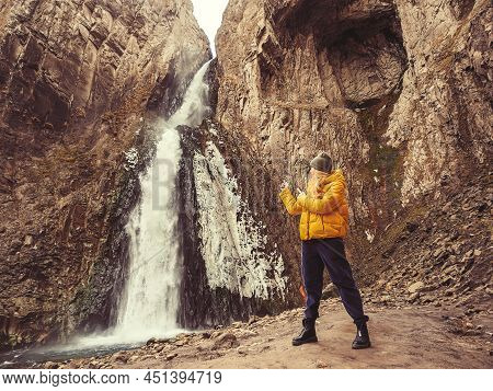 A Woman Points Her Fingers At An Icy Powerful Waterfall Flowing From A Gorge In The Sheer Cliffs Of 