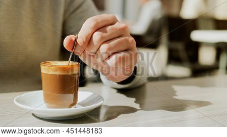 a young caucasian man stirs his machiatto, served in a glass, with a coffee spoon sitting at a table of a sidewalk cafe