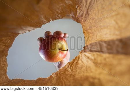 A Hand Takes A Red Apple From A Brown Paper Bag. A Mature Man's Hand Holds The Ripe Fruit Over The O