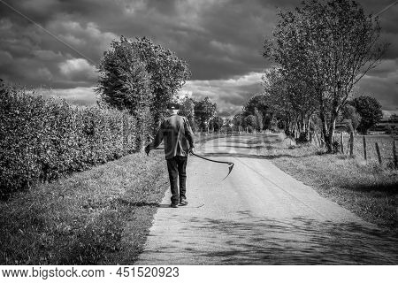 Bullingen, Walloon Regio - East Belgium - 08 27 2019 An Old Farmer Carrying A Scythe, Looking Like T