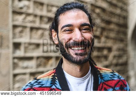 Merry Hispanic Man In Colorful Jacket With Beard And Ponytail Smiling And Looking At Camera. While S