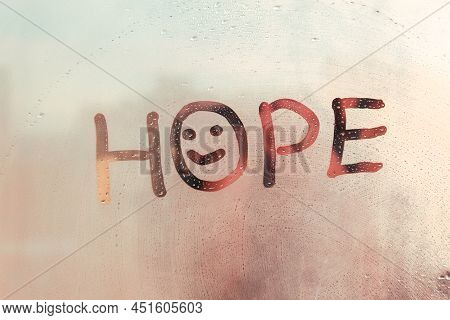 Handwritten Word Hope With Doodle Smile On Misted Sunset Glass On Window Flooded With Raindrops
