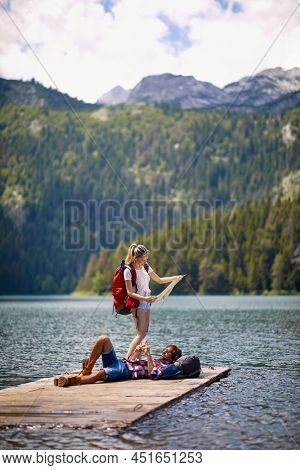 Young couple with bacpacks on adventure. Man lying on wooden jetty by lake, woman looking at map. Lifestyle, togetherness, nature concept