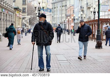 Moscow, Russia - March 2022: Elderly Man In Lowered Medical Mask Walking With A Cane On A City Stree