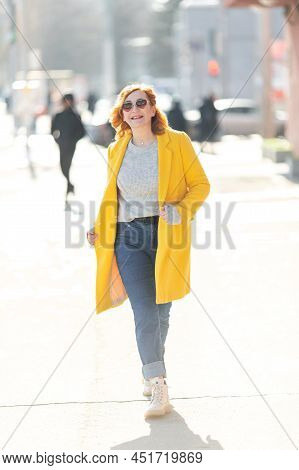 A Young Active Woman With Red Hair, In Bright Clothes, A Yellow Coat And Jeans Is Walking Around The