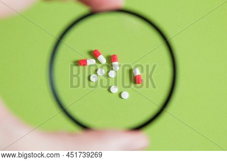 A few pills and capsules through a magnifying glass is a table. A black filter surrounds white and red medications on a green background.