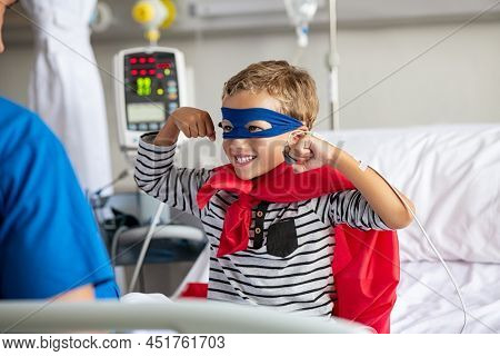 Cheerful strong little boy wearing blue eyeband and red cape like superhero playing with nurse. Playful child gesturing dressed in superhero costume at clinic overcome adversity and health challenge. 
