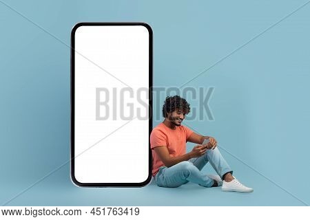 Rlaxed Young Indian Man Using Cell Phone, Sitting On Blue