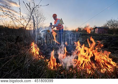 Fireman Ecologist Extinguishing Fire In Field With Evening Sky On Background. Male Environmentalist 