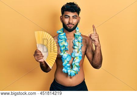 Arab man with beard wearing hawaiian lei holding handfan smiling with an idea or question pointing finger with happy face, number one 