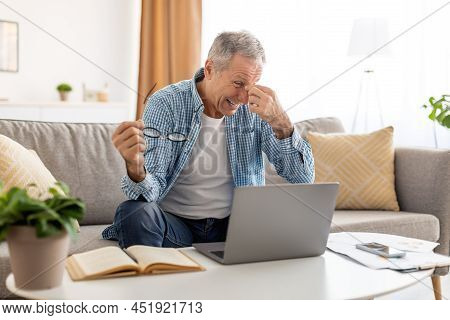 Tired Mature Man Sitting At Desk With Pc Massaging Nosebridge