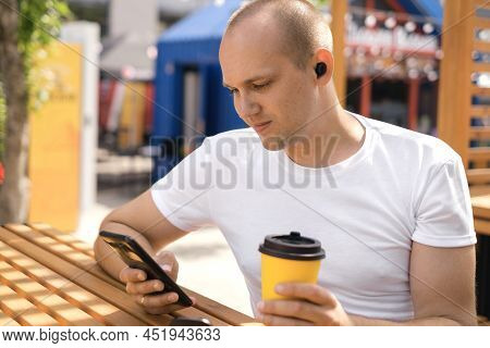 A Man Is Sitting At A Table In A Cafe Drinking Coffee From A Disposable Cup, Listening To Music Thro