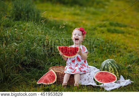 Little Girl In A White Dress Eats A Watermelon At A Picnic. She Laughs Merrily Sitting On A Wicker B