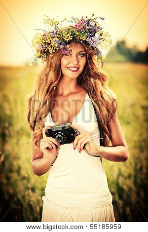 Portrait of a romantic smiling young woman in a circlet of flowers standing with her old camera outdoors.