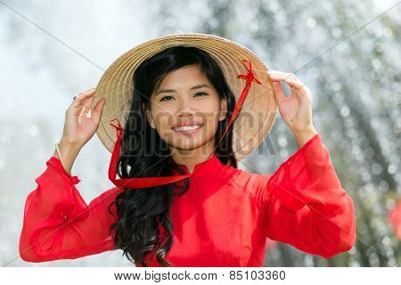 Smiling vivacious Vietnamese woman in a traditional red outfit and conical hat standing in front of a fountain smiling at the camera